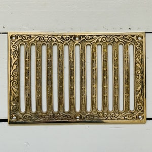 May include: A gold-toned metal vent cover with a decorative, ornate design. The cover has a series of vertical slots for air circulation.