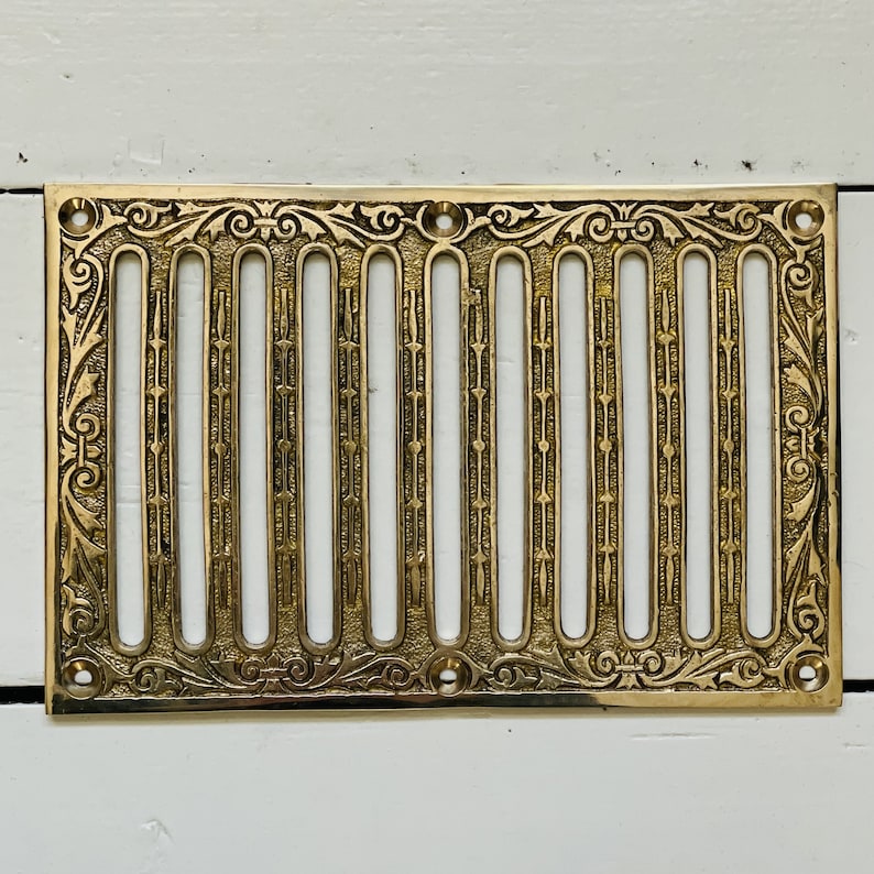 May include: A gold-toned metal vent cover with a decorative, ornate design. The cover has a series of vertical slots for air circulation.