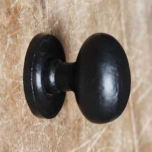 May include: Close-up of a black, oval-shaped door knob with a circular base. The knob is mounted on a light brown, textured wooden surface. The hardware has a matte finish.