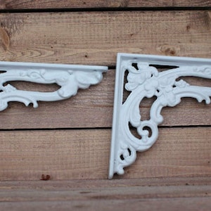May include: Two white metal shelf brackets with a decorative floral design. The brackets are in the shape of a right angle and have a scalloped edge.