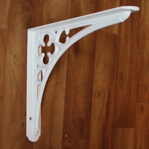 Puede incluir: Soporte de estante de metal blanco con un dise&ntilde;o decorativo y ornamentado. El soporte tiene forma de &aacute;ngulo recto y un borde festoneado.