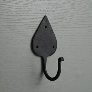 Puede incluir: Un gancho de pared negro con forma de hoja, hecho de metal. El gancho tiene una parte superior en forma de lágrima con tres agujeros para montaje y un gancho curvado. Montado sobre una superficie de madera gris.