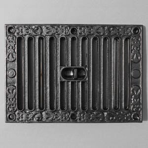 May include: Black cast iron vent cover with a decorative floral design around the perimeter. The vent cover has a series of vertical bars with a small rectangular opening in the center.