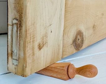 May include: A wooden doorstop with a wedge shape and a handle. The doorstop is made of light brown wood and has a natural finish.