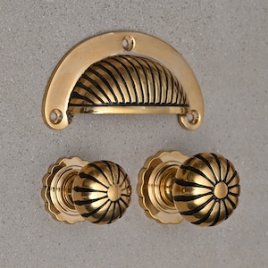 May include: A set of three gold-toned cabinet hardware pieces. The set includes a decorative drawer pull and two round knobs. The drawer pull has a ribbed design and the knobs have a sunburst design.