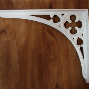 Puede incluir: Soporte de estante de hierro fundido blanco con un dise&ntilde;o decorativo y ornamentado. El soporte tiene una parte superior curva y una parte inferior puntiaguda.