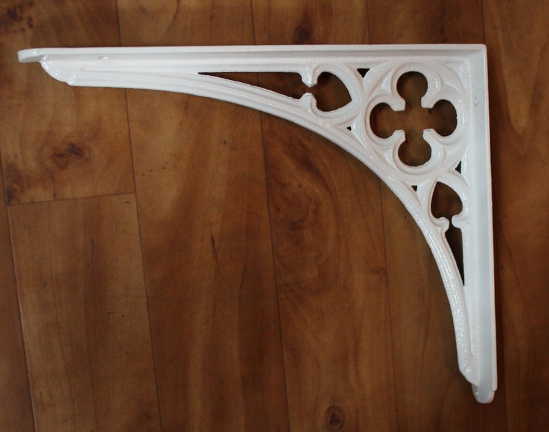 Puede incluir: Soporte de estante de hierro fundido blanco con un dise&ntilde;o decorativo y ornamentado. El soporte tiene una parte superior curva y una parte inferior puntiaguda.
