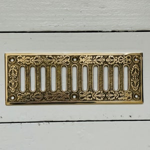 May include: A gold-colored metal vent cover with a decorative design of leaves and swirls. The vent cover has 12 rectangular openings.