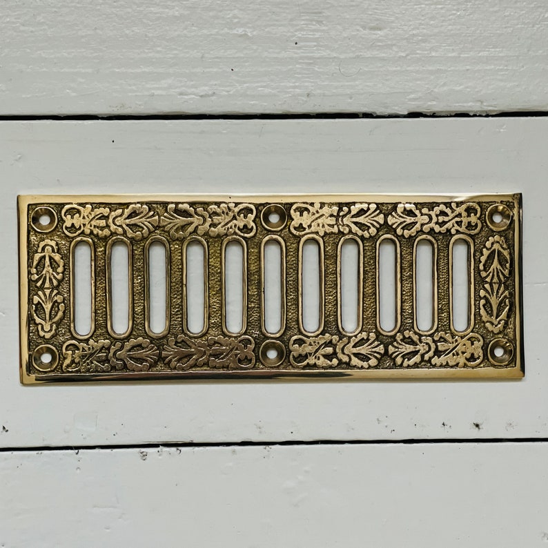 May include: A gold-colored metal vent cover with a decorative design of leaves and swirls. The vent cover has 12 rectangular openings.