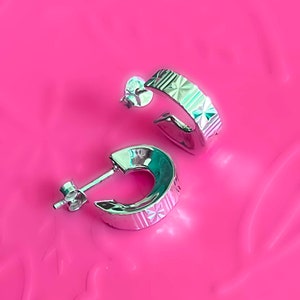 May include: A pair of silver hoop earrings with a textured, geometric design on the outer band. The earrings are set against a bright pink background, showcasing their polished, reflective surface.