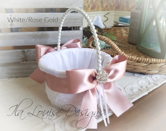May include: White and rose gold flower girl basket with a pearl handle and a large satin bow. The basket has a white lace exterior and a white fabric lining. The bow is adorned with a rhinestone embellishment. The text "White/Rose Gold" is visible.