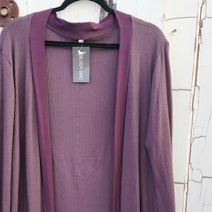 May include: A long, open-front cardigan in shades of purple, with ribbed texture and two front pockets. The cardigan has a contrasting dark purple trim on the collar, cuffs, and pockets. A tag with text hangs from the collar.
