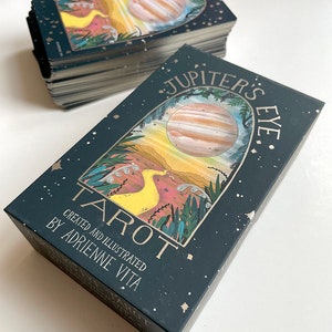 May include: A deck of Jupiter's Eye Tarot cards, featuring a dark green box with gold lettering and a colorful illustration of a planet and a yellow bird. The box reads "Jupiter's Eye Tarot" and "Created and Illustrated by Adrienne Vita".