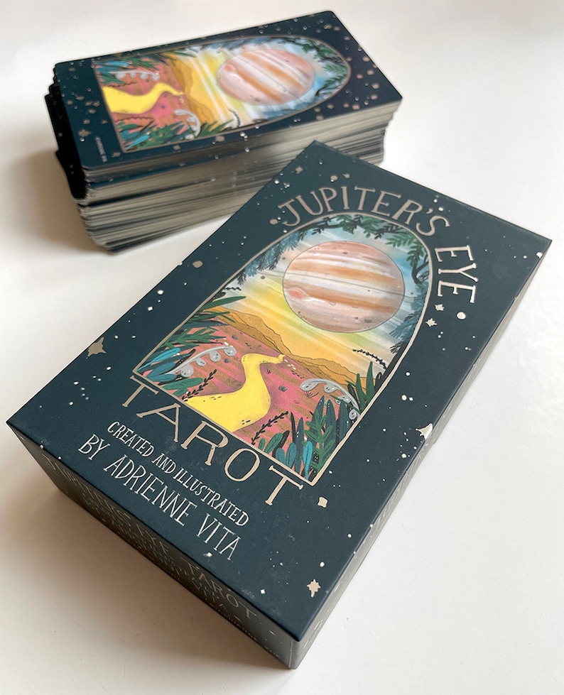 May include: A deck of Jupiter's Eye Tarot cards, featuring a dark green box with gold lettering and a colorful illustration of a planet and a yellow bird. The box reads "Jupiter's Eye Tarot" and "Created and Illustrated by Adrienne Vita".