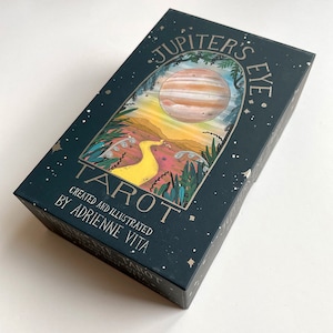 May include: A dark green box with gold lettering that reads "Jupiter's Eye Tarot" and "Created and Illustrated by Adrienne Vita". The box features a colorful illustration of a planet with rings, a yellow path, and green foliage.