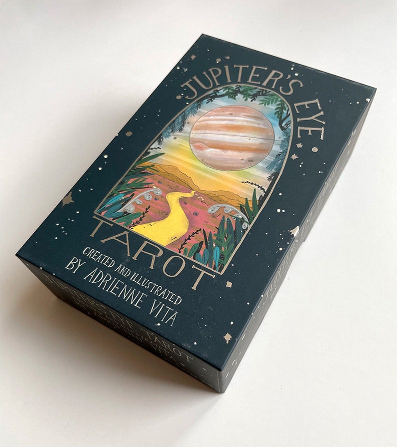 May include: A dark green box with gold lettering that reads "Jupiter's Eye Tarot" and "Created and Illustrated by Adrienne Vita". The box features a colorful illustration of a planet with rings, a yellow path, and green foliage.