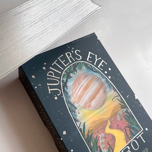 May include: A box of Jupiter's Eye Tarot cards. The box is dark blue with gold lettering and a colorful illustration of a planet, a yellow path, and green plants. The text on the box reads "Jupiter's Eye Tarot. Created and illustrated by DRIENNE VITTA."