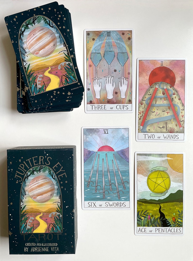 May include: A deck of tarot cards with a dark blue and gold box featuring a stylized illustration of Jupiter. The cards are illustrated with colorful scenes and feature the text "Jupiter's Eye Tarot" and "Created and Illustrated by Adrienne Vita".