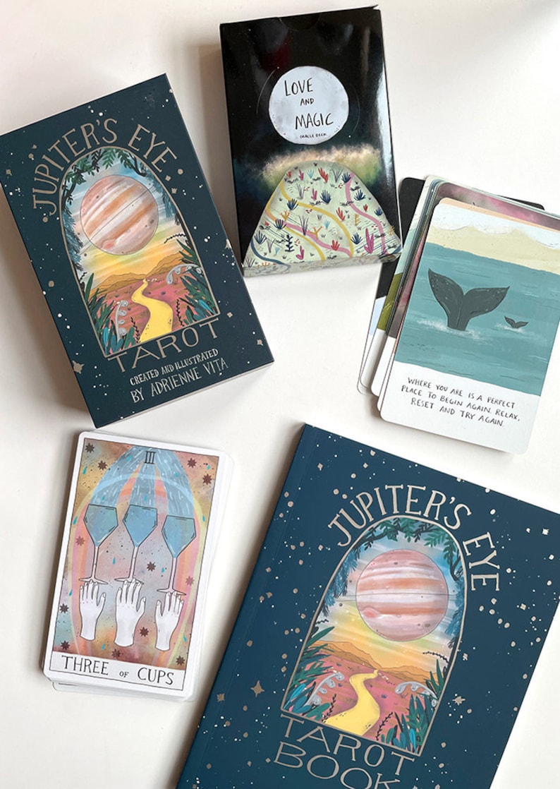 May include: A tarot deck with a teal blue background and a gold inscription that reads "Jupiter's Eye Tarot". The deck features a watercolor illustration of a planet with a yellow path leading to it. The card in the foreground is the Three of Cups card, featuring three cups with water pouring out of them and three hands reaching up to the cups.