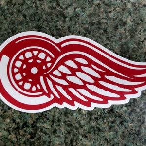 May include: Red and white Detroit Red Wings logo sticker. The logo features a stylized wing with a wheel inside.