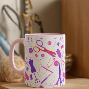 Peut inclure: Une tasse en céramique blanche avec un bord rose présente un motif coloré d'objets liés à la couture, notamment des ciseaux, des aiguilles, des bobines de fil, des boutons et un mannequin de couture.