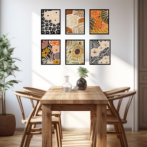 May include: Six framed prints with abstract floral and leaf designs in shades of orange, yellow, brown, and black. The prints are hung on a white wall above a wooden dining table with four chairs.