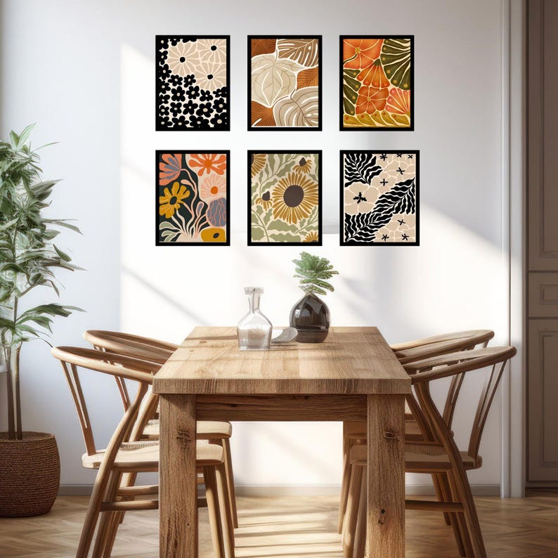 May include: Six framed prints with abstract floral and leaf designs in shades of orange, yellow, brown, and black. The prints are hung on a white wall above a wooden dining table with four chairs.