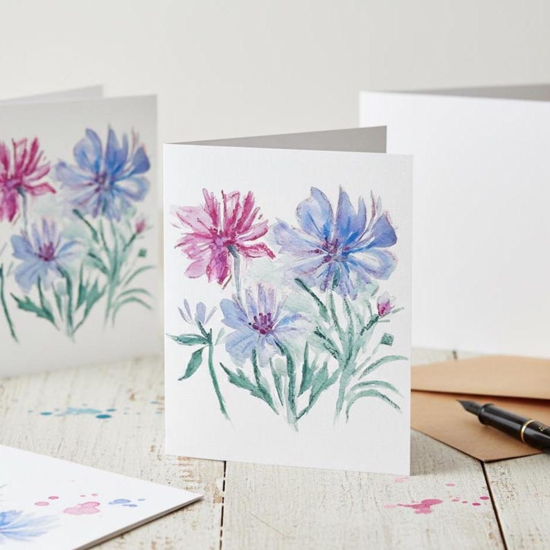 Puede incluir: Tarjetas de felicitaci&oacute;n florales en acuarela con flores rosas y azules con tallos y hojas verdes. Las tarjetas est&aacute;n sobre un fondo blanco, con una superficie de madera. Tambi&eacute;n se ven un bol&iacute;grafo y un sobre.