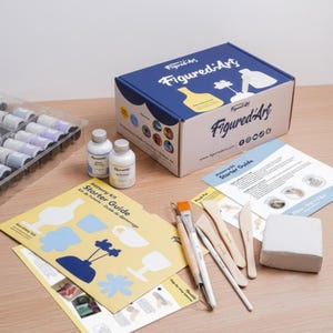 May include: A pottery kit with a blue and white box labeled "FiguredArt" and various art supplies. The kit includes paint bottles, tools, a starter guide, and a block of clay. The box has a logo with a vase and flower.