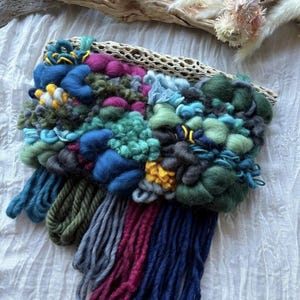 Może przedstawiać: Kolorowy, tkany wiszący na ścianę z różnymi fakturami i kolorami, w tym niebieskim, zielonym, czerwonym i żółtym. Wisiorek jest wykonany z przędzy i ma frędzle u dołu.