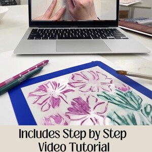 Puede incluir: Un port&aacute;til muestra un tutorial en v&iacute;deo. Debajo, una alfombrilla azul contiene una pintura floral con acentos morados y verdes. Tambi&eacute;n se ven dos rotuladores y un pincel. El texto "Includes Step by Step Video Tutorial" est&aacute; en la parte inferior.