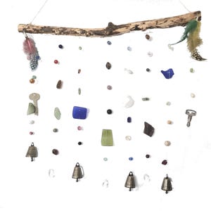 May include: A rustic wall hanging featuring a natural wooden branch with hanging strands of beads, glass pieces, keys, and small brass bells. Colorful feathers adorn each end of the branch, creating a unique, bohemian-style decor.