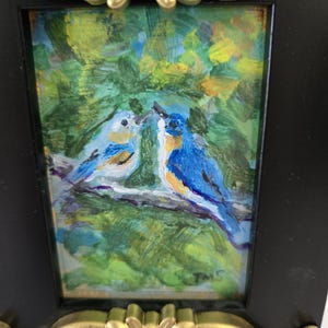 May include: A small rectangular painting of two blue birds perched on a branch against a green background, encased in a black frame with ornate gold-colored decorative accents at the top and bottom. The artist's initials TMS are visible.