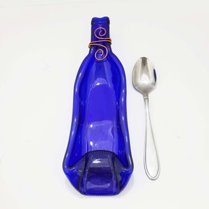 May include: A cobalt blue, flattened glass bottle shaped into a serving tray. The bottle's neck is adorned with copper wire swirls. A stainless steel spoon rests beside the tray on a white surface. The tray is likely for serving appetizers.