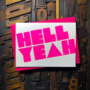 May include: A white card with the words "HELL YEAH" printed in bright pink letters. The card is sitting on a background of wooden printing blocks.