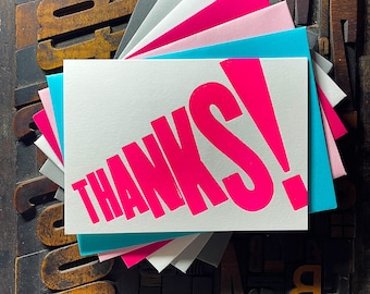May include: A stack of blank greeting cards with a pink "THANKS!" card on top. The cards are arranged in a fan shape and are sitting on a wooden surface with old printing blocks.