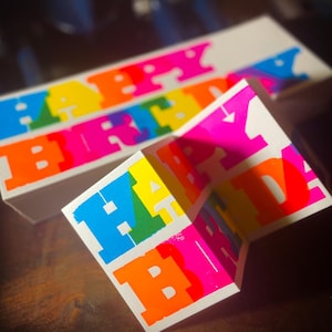 May include: A white card featuring the words "HAPPY BIRTHDAY" in large, colourful letters. The letters are in a rainbow of colours, including blue, green, yellow, orange, and pink. The card is folded in an accordion style.
