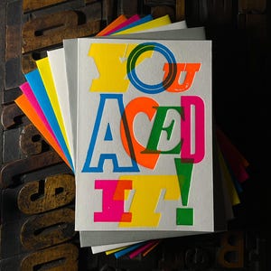 May include: A set of greeting cards featuring the text "YOU ACED IT!" in vibrant, overlapping colours. The cards are white with a fanned-out arrangement, revealing the edges of the colourful paper. The background is a dark wooden surface.