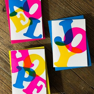 May include: Three greeting cards with the words "NOEL", "JOY", and "HOPE" printed in overlapping, vibrant colors. Each card has a different colored backing: pink, blue, and yellow. The cards are on a wooden surface.