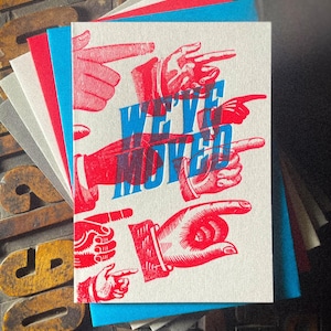 May include: A stack of greeting cards with a design featuring red and blue hands pointing in various directions. The text "WE'VE MOVED" is printed in blue and red. The cards have a white background and are stacked with red, blue, and grey cardstock.