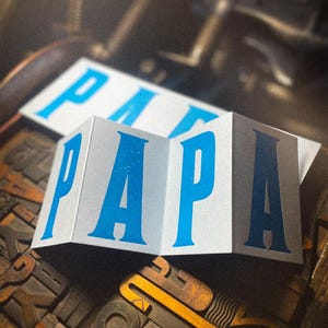 May include: A folded card displaying the word "PAPA" in bold, light blue lettering. The card has a white, silvery finish and rests on a wooden surface with vintage printing blocks. The word "PAPA" is also printed on a flat card in the background.