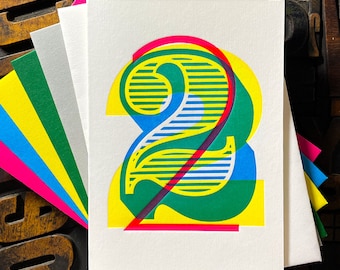 Puede incluir: Una tarjeta de felicitación blanca con un número dos grande y estilizado, impreso en capas superpuestas de tintas amarillo vibrante, azul cian, rosa magenta y verde. El diseño incorpora patrones de líneas horizontales en el número.