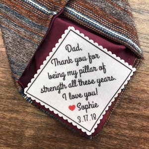 May include: A white tag with scalloped edges attached to a brown and blue striped tie. The tag has a handwritten message that reads "Dad, Thank you for being my pillar of strength all these years. I love you! Sophie 3.17.18".