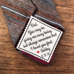 May include: A patterned necktie with a personalized message. The tie features a mix of brown, blue, and white stripes. A white, scalloped-edged patch is sewn into the tie, with the text "Dad, You may be giving me away today, but always remember that I loved you first. Ashley 6.15.19."