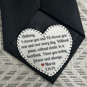 May include: A white heart-shaped tag with a black stitched border and a message that reads "Anthony, I choose you and I'll choose you over and over every day. Without pause, without doubt, in a heartbeat. I love you today, forever and always. Marcie 2.14.24". A small red heart is next to the name Marcie.