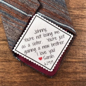 May include: A white card with a handwritten message on it. The message reads "Johnny, You're not losing me as a sister. You're just gaining a new brother. I love you! Sarah 6.29.19". The card is attached to a brown and blue striped tie.