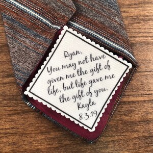 May include: A white card with scalloped edges and a handwritten message that reads "Ryan, You may not have given me the gift of life, but life gave me the gift of you. Kayla 8.3.19". The card is attached to a brown and blue striped tie.