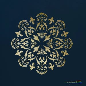 Op de afbeelding: Gouden stencil met een gedetailleerd bloemenontwerp en bijenmotieven op een donkerblauwe achtergrond. De stencil is rond en heeft een delicaat, ingewikkeld patroon.