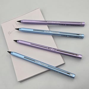 Peut inclure: Quatre crayons violets et bleus, avec des pointes noires, reposent sur un bloc-notes blanc. Les crayons portent des inscriptions telles que "NCC-1701" et "U.S.S. Enterprise". Le bloc-notes porte l'inscription "Pencil!"