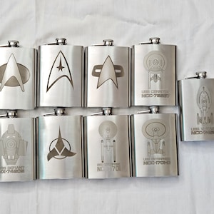 Könnte beinhalten: Acht Edelstahl-Schnapsflaschen mit Star Trek-Schiffsemblemen und -Namen. Die Flaschen zeigen die USS Enterprise, USS Defiant, USS Voyager, USS Cerritos und das Starfleet-Emblem.